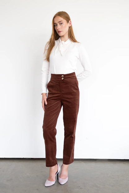 A woman wearing the Graham Corduroy Pants by Eve Lavoie in Hazelnut, with a high waist, double button, slim fit and ankle length leg. She is wearing them with a white top and standing in front of a white background. 