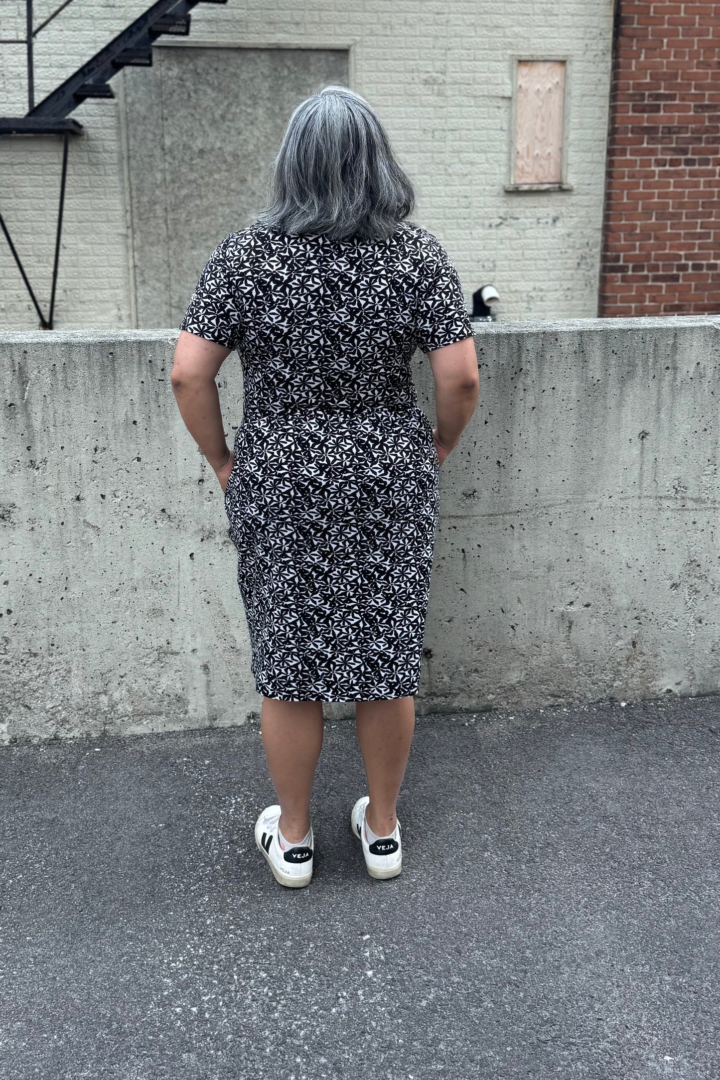 Back view of a woman wearing the Hayley Dress by Tangente in Black and White floral print, a short sleeved bamboo dress with a gathered knee-length skirt. She is standing outside in front of a cement wall. 