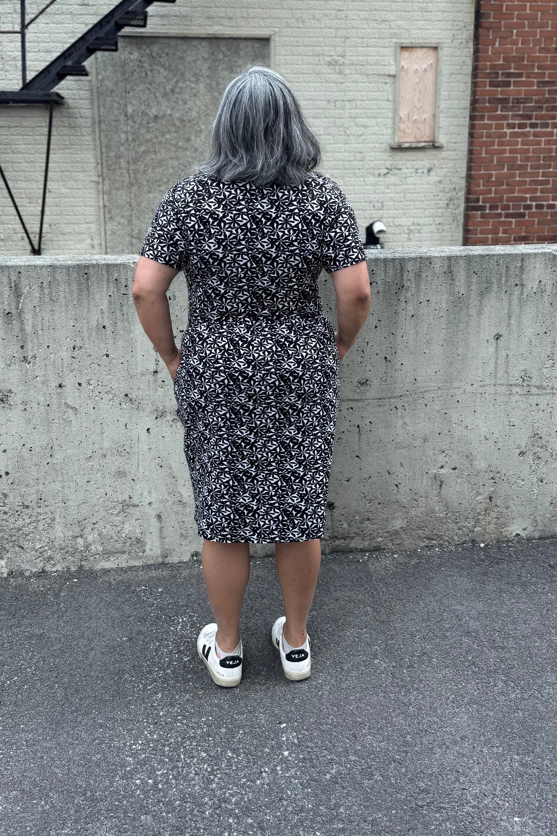 Back view of a woman wearing the Hayley Dress by Tangente in Black and White floral print, a short sleeved bamboo dress with a gathered knee-length skirt. She is standing outside in front of a cement wall. 