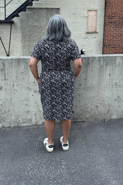 Back view of a woman wearing the Hayley Dress by Tangente in Black and White floral print, a short sleeved bamboo dress with a gathered knee-length skirt. She is standing outside in front of a cement wall. 