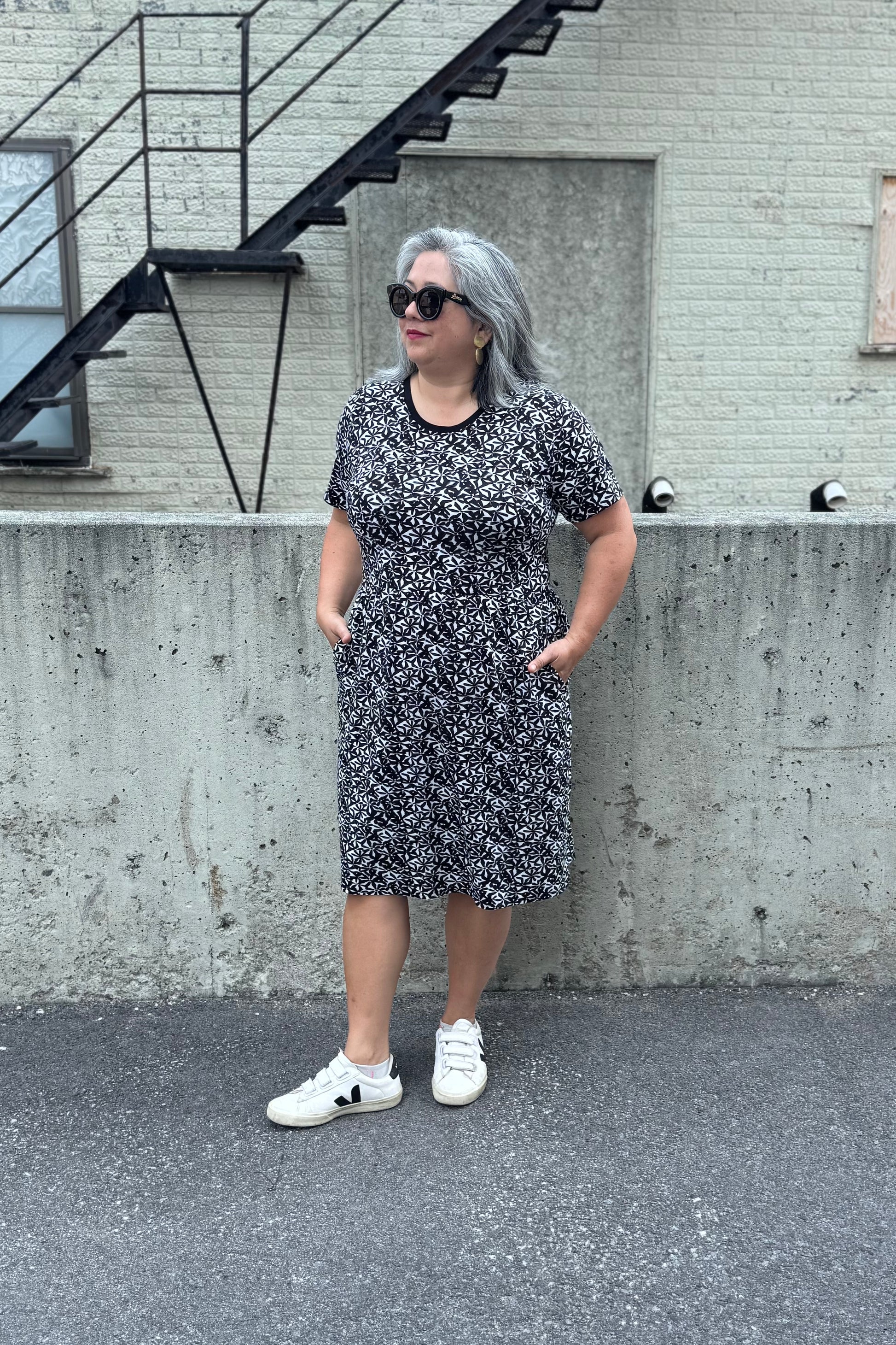 A woman wearing the Hayley Dress by Tangente in Black and White floral print, a short sleeved bamboo dress with a gathered knee-length skirt. She is standing outside in front of a cement wall. 