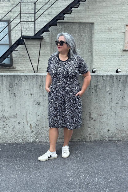 A woman wearing the Hayley Dress by Tangente in Black and White floral print, a short sleeved bamboo dress with a gathered knee-length skirt. She is standing outside in front of a cement wall. 