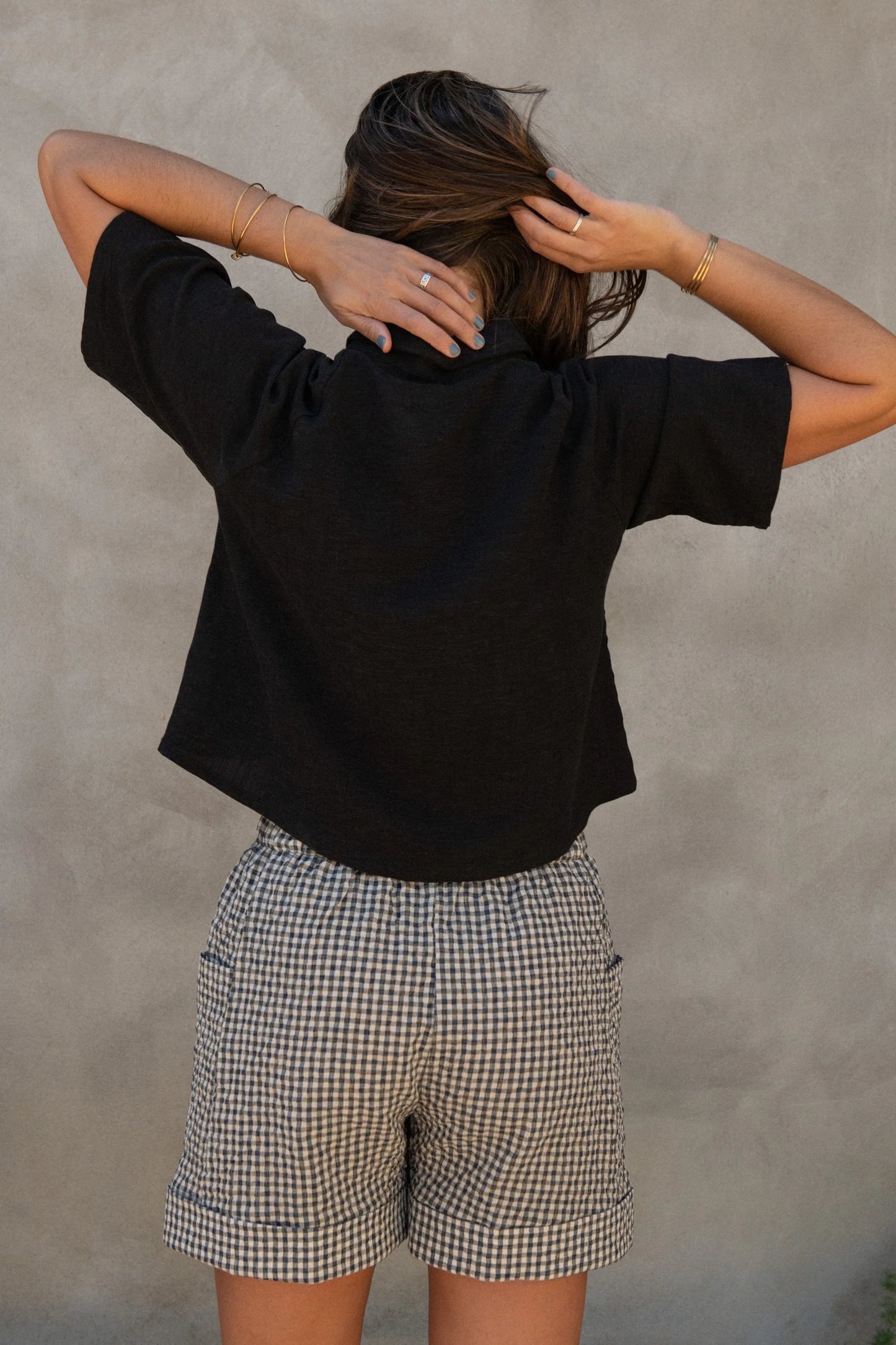Back view of a woman wearing the Hosta Screen-printed Blouse by Kazak in Black with Dandelions, with hand-screen-printed flowers, mid-length raglan sleeves, a classic collar, and a button front. She is wearing it with gingham shorts and standing outside by a grey wall. 