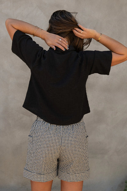 Back view of a woman wearing the Hosta Screen-printed Blouse by Kazak in Black with Dandelions, with hand-screen-printed flowers, mid-length raglan sleeves, a classic collar, and a button front. She is wearing it with gingham shorts and standing outside by a grey wall. 