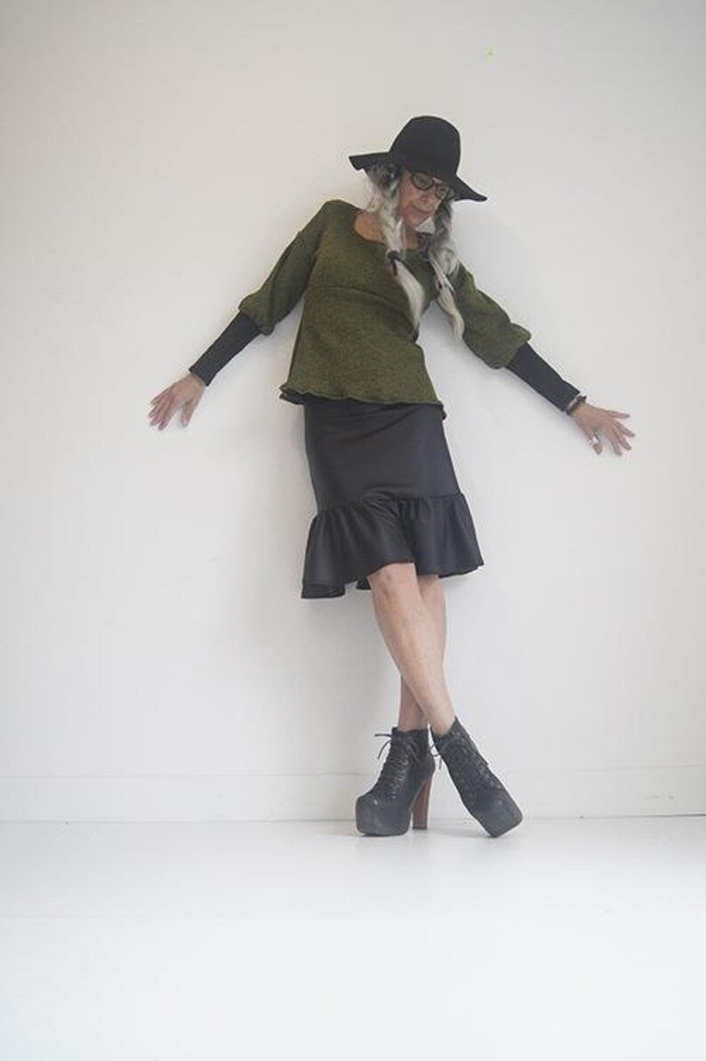 A woman wearing the Athena Top by SI Design in Olive Green, with a square neckline, an empire waist, and bishop style sleeves with long cuffs in black lace. She is wearing it with a black skirt and is standing in front of a white background 