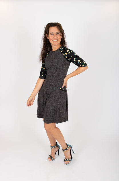 A woman wearing the Raphaelle Dress from Infime in Black, an A-line knee-length dress with 3/4 raglan sleeves that feature a floral print and pockets with a floral detail. She is standing in front of a white background