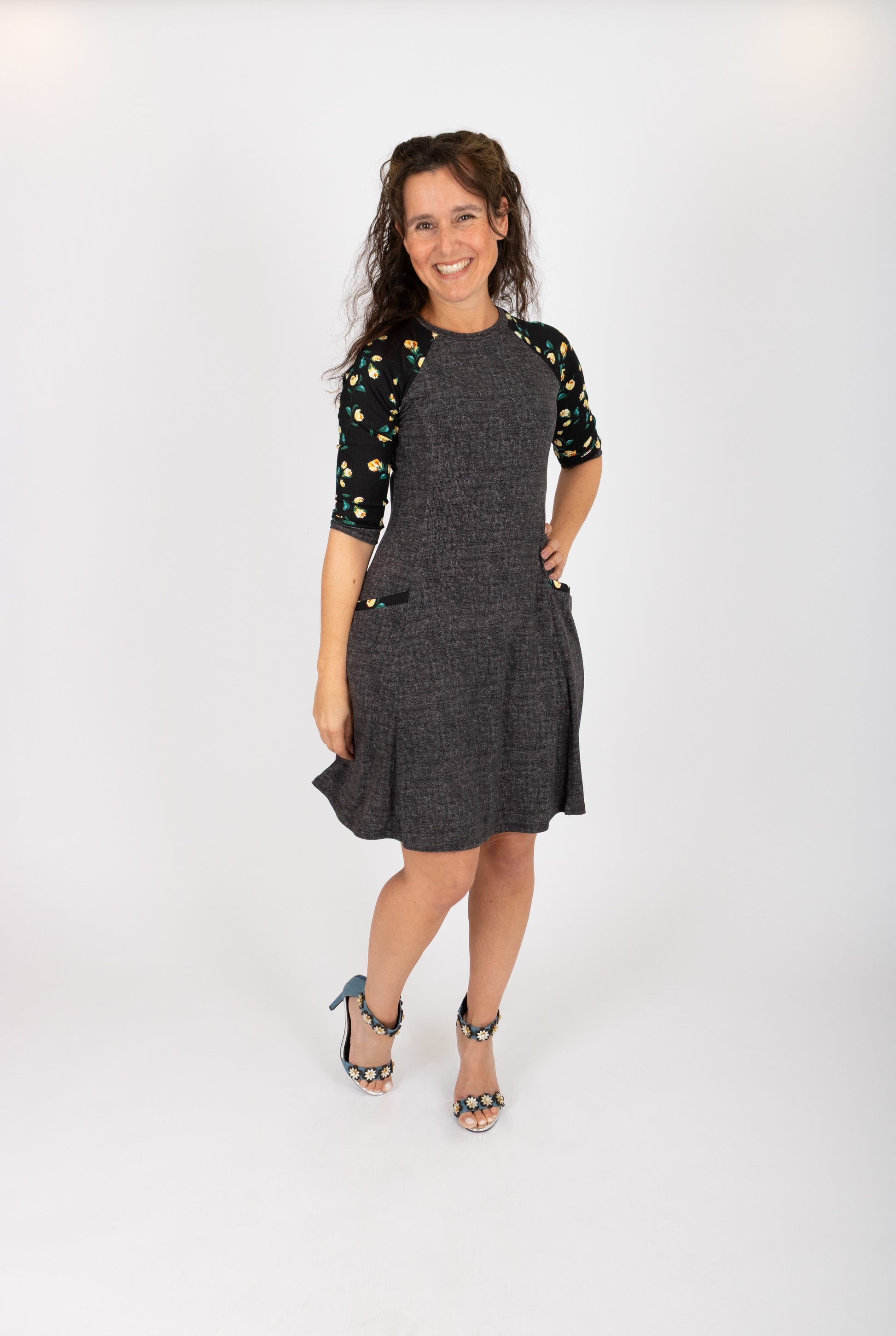A woman wearing the Raphaelle Dress from Infime in Black, an A-line knee-length dress with 3/4 raglan sleeves that feature a floral print and pockets with a floral detail. She is standing in front of a white background