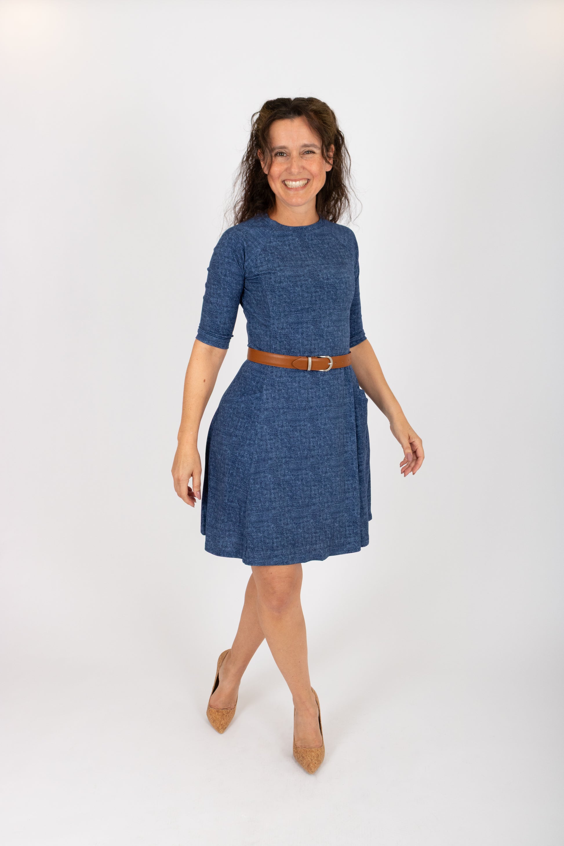 A woman wearing the Raphaelle Dress from Infime in Blue, an A-line knee-length dress with 3/4 raglan sleeves and pockets. She is standing in front of a white background.