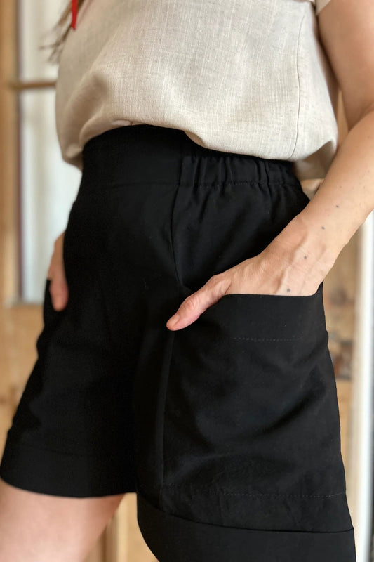 Waist down view of a woman wearing the Irish Shorts by Kazak in Black, with a smooth front waist, elastic at the back, large pockets, and mid-thigh length. She is wearing them with a cream top and standing in a room with a door. 
