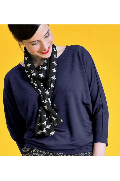 Waist-up view of a woman wearing the Jo Slouch Tee by Mandala in Cadet with a scarf, standing in front of a yellow background 