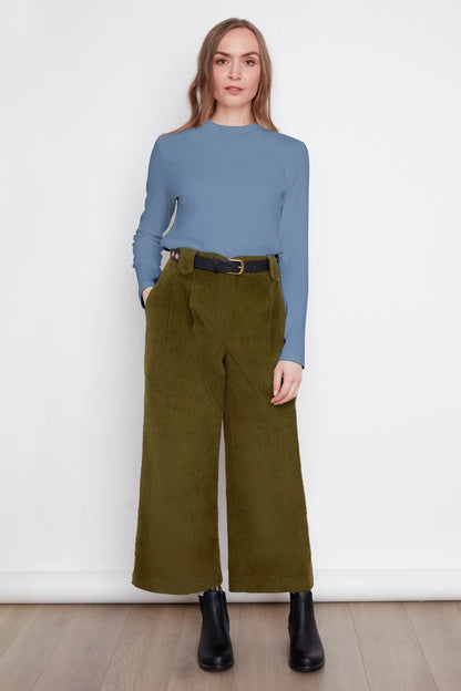 A woman wearing the Idadora Pants by Julei in Moss, wide-leg corduroy pants. She is wearing them with a blue sweater and standing against a white background