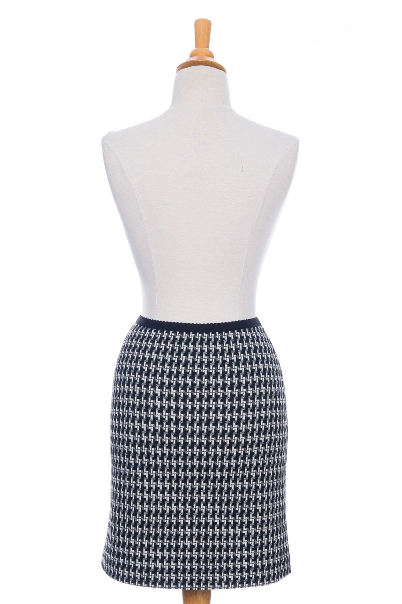 A back view of the Meleze Skirt by Rien ne se Perd in Black and White houndstooth, a knee-length straight skirt with an elastic waist, is shown on a mannequin in front of a white background. 