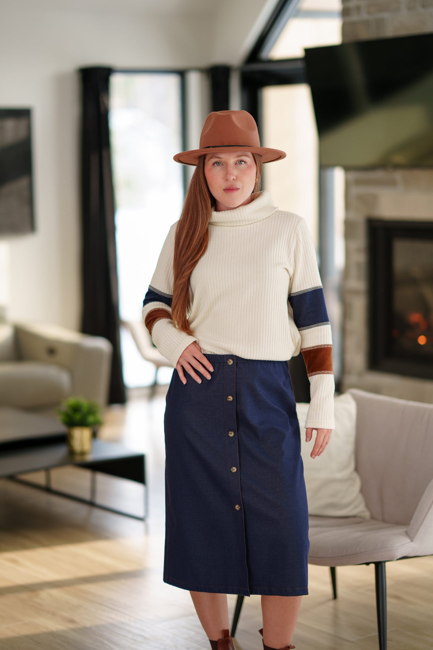 A woman wearing the Nicolet Skirt from Rien ne se Perd, a blue denim midi with buttons up the front, an elasticated waist, and a centre back slit. She is wearing it with a colour blocked sweater and a brown hat and standing in a living room. 