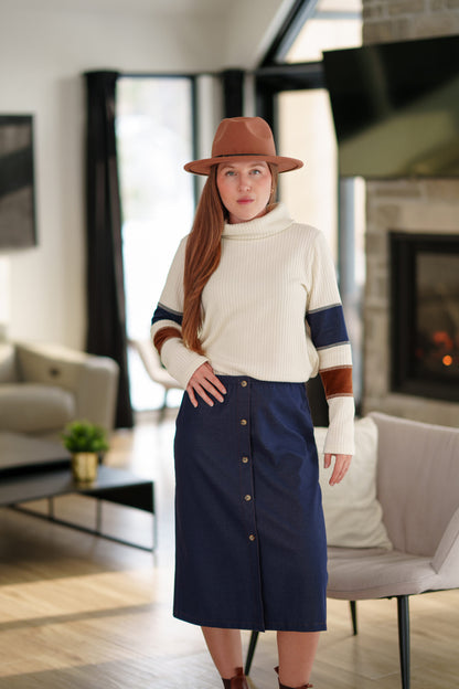 A woman wearing the Nicolet Skirt from Rien ne se Perd, a blue denim midi with buttons up the front, an elasticated waist, and a centre back slit. She is wearing it with a colour blocked sweater and a brown hat and standing in a living room. 
