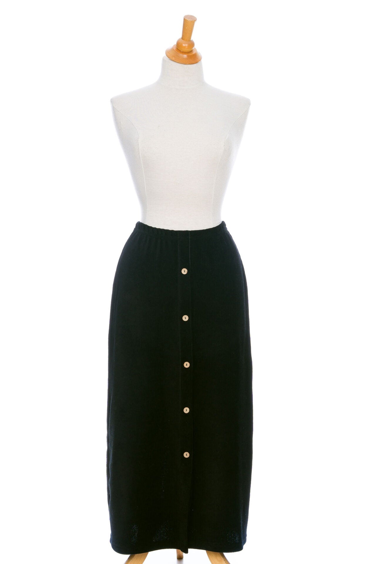 The Pruche Skirt by Rien ne se Perd, a 7/8 length Black corduroy skirt with an elastic waist, five coconut buttons up the front, and a slit at the back, is shown on a mannequin against a white background. 