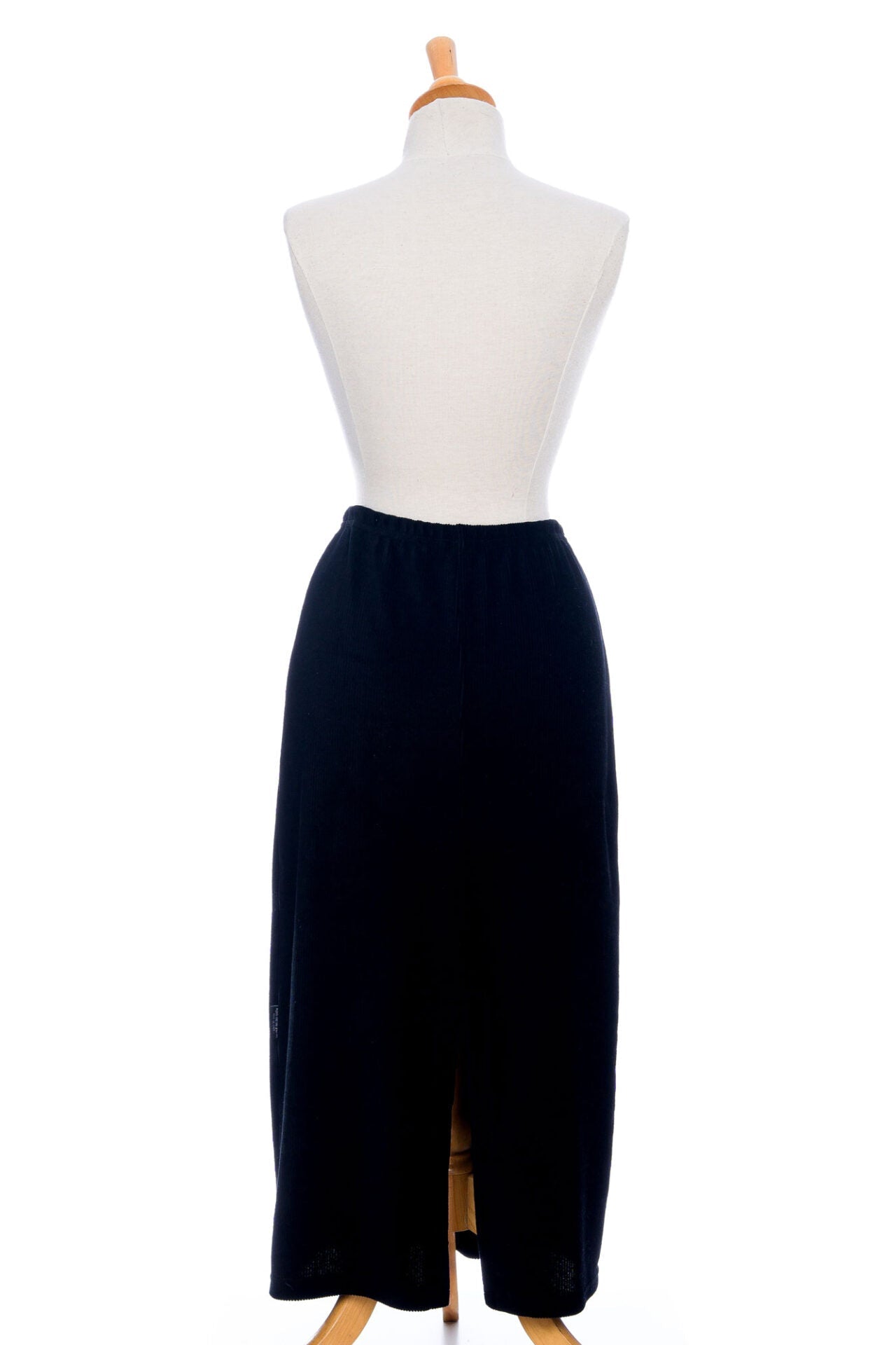 A back view of the Pruche Skirt by Rien ne se Perd, a 7/8 length Black corduroy skirt with an elastic waist, five coconut buttons up the front, and a slit at the back, is shown on a mannequin against a white background. 