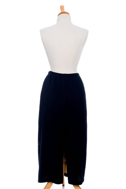 A back view of the Pruche Skirt by Rien ne se Perd, a 7/8 length Black corduroy skirt with an elastic waist, five coconut buttons up the front, and a slit at the back, is shown on a mannequin against a white background. 