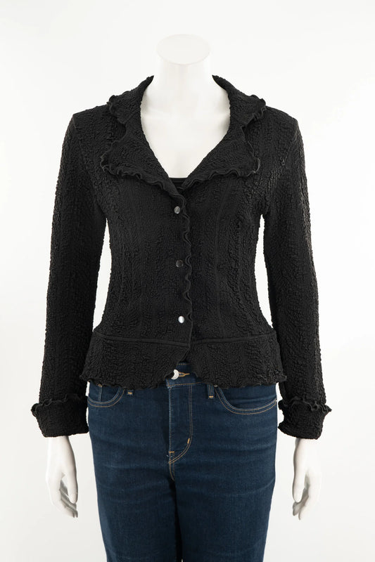 The Essential Jacket - Jasper by Karkass is shown on a mannequin in front of a white background. It's a black textured jacket with a ruffled collar, button closure, and frill details at the cuffs and hem. 