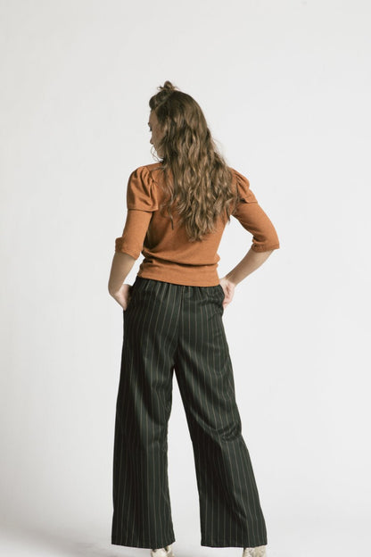 Back view of a woman wearing the Katherine Pants by Allison Wonderland in Pinstripe, with the Eyre Sweater in Caramel, standing in front of a white background 