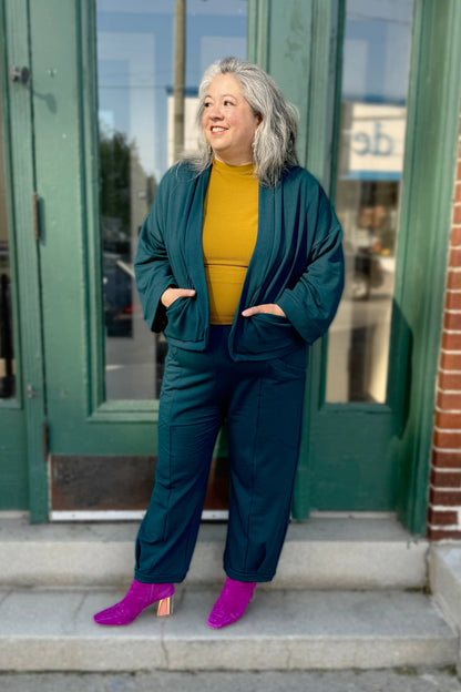 Christina wearing the Herbst Pant and Thuya Jacket by Kazak in Teal, standing in front of a green door 