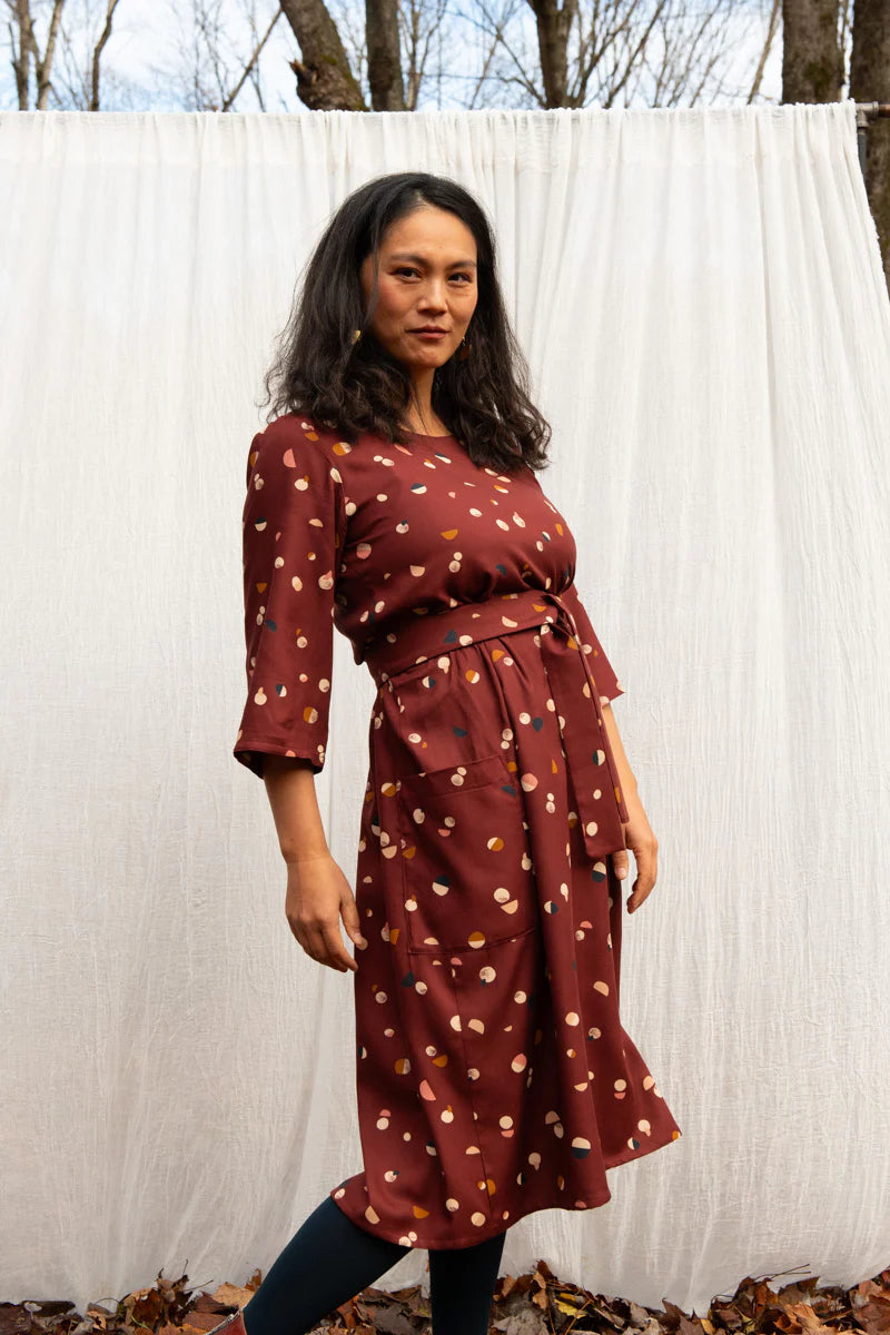 A woman wearing the Persea Dress by Kazak in Bordeaux Moon print, a below-the-knee length dress with 3/4 sleeves, a removable belt, and large patch pockets. She is standing in a forest in front of a white sheet. 