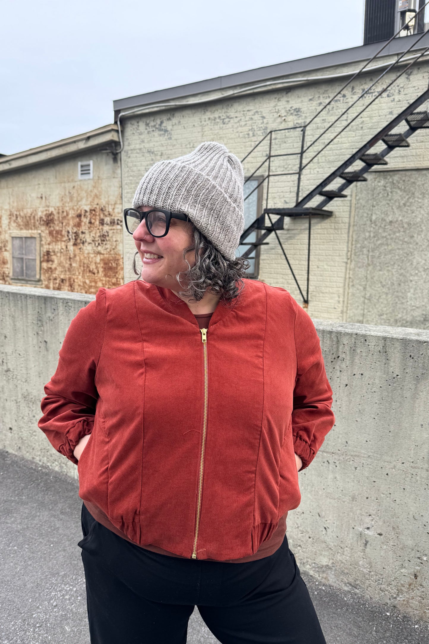 A woman wearing the Ribbed Beanie by Charlevoix Pure Laine in Grey. She is wearing it with the Redrawn Zip Jacket by Eve Lavoie and is standing outside. 