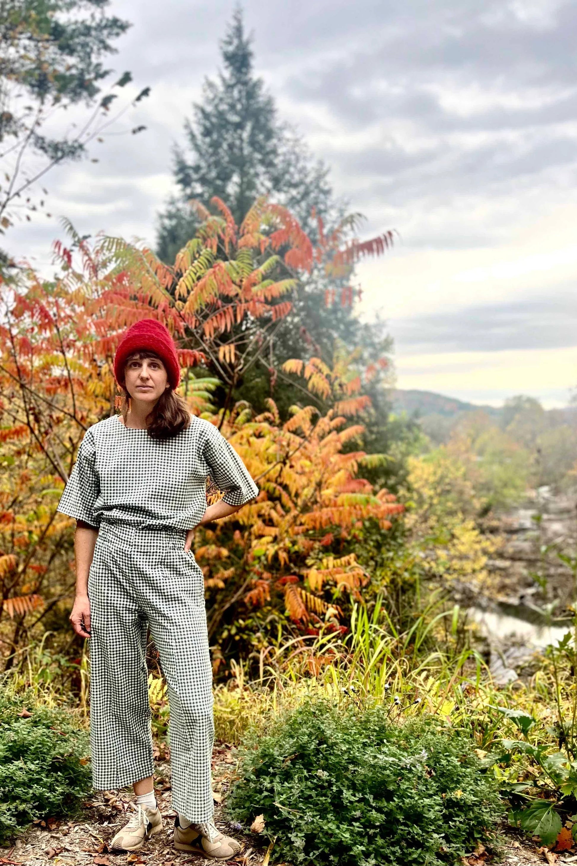 A woman wearing the Olea Pants by 
Kazak in Forest Vichy, wide-legged ankle-length pants with a flat-front elastic waist and large patch pockets. She is wearing them with the matching Abelia Top and standing in a forest.