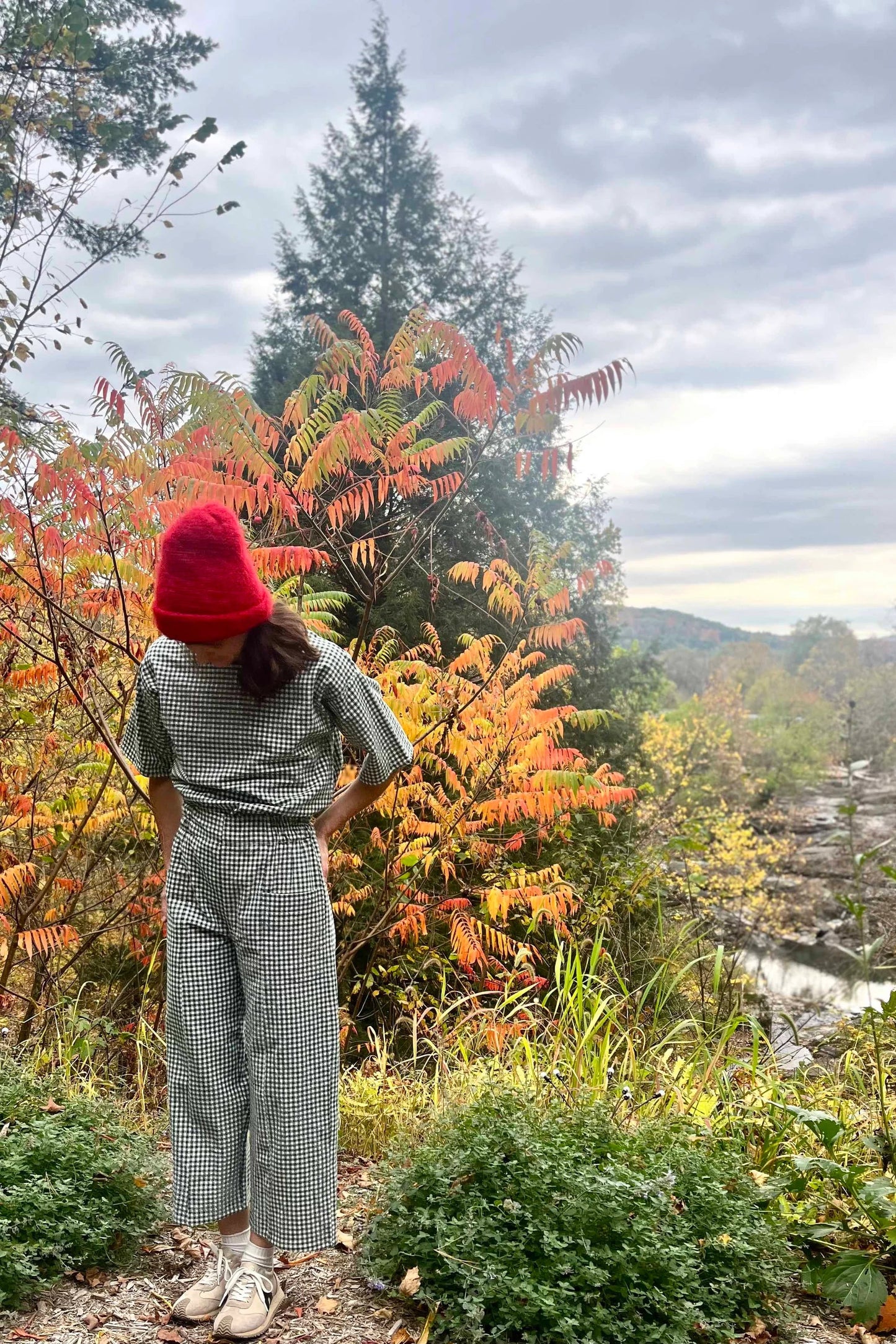 A woman wearing the Olea Pants by 
Kazak in Forest Vichy, wide-legged ankle-length pants with a flat-front elastic waist and large patch pockets. She is wearing them with the matching Abelia Top and standing in a forest.