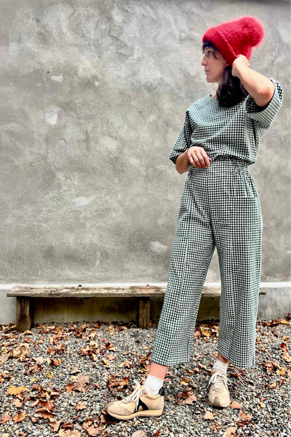 A woman wearing the Olea Pants by 
Kazak in Forest Vichy, wide-legged ankle-length pants with a flat-front elastic waist and large patch pockets. She is wearing them with the matching Abelia Top and standing outside by a grey wall. 