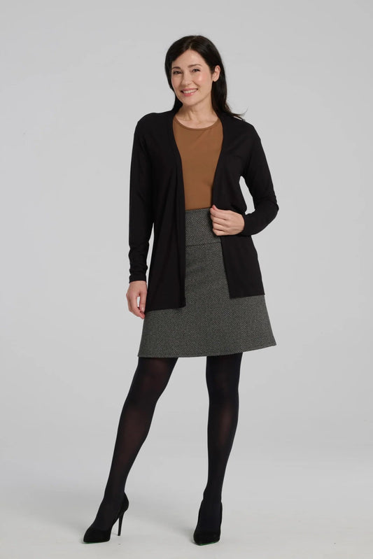 A woman wearing the Frost Skirt by Kollontai in Charcoal tweed, an above the knee length A-line skirt with an elastic waist.  She is wearing it with a brown top and a black cardigan and standing in front of a grey background. 