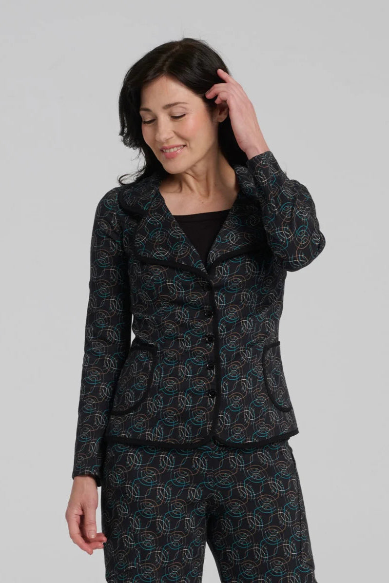 A woman wearing the Nara Jacket by Kollontai in Turquoise, a blazer with a turquoise on black geometric print, wit a rounded lapel collar, slim fit, and side pockets. She is wearing it with a black top and the matching Elma Pant and standing in front of a grey background. 