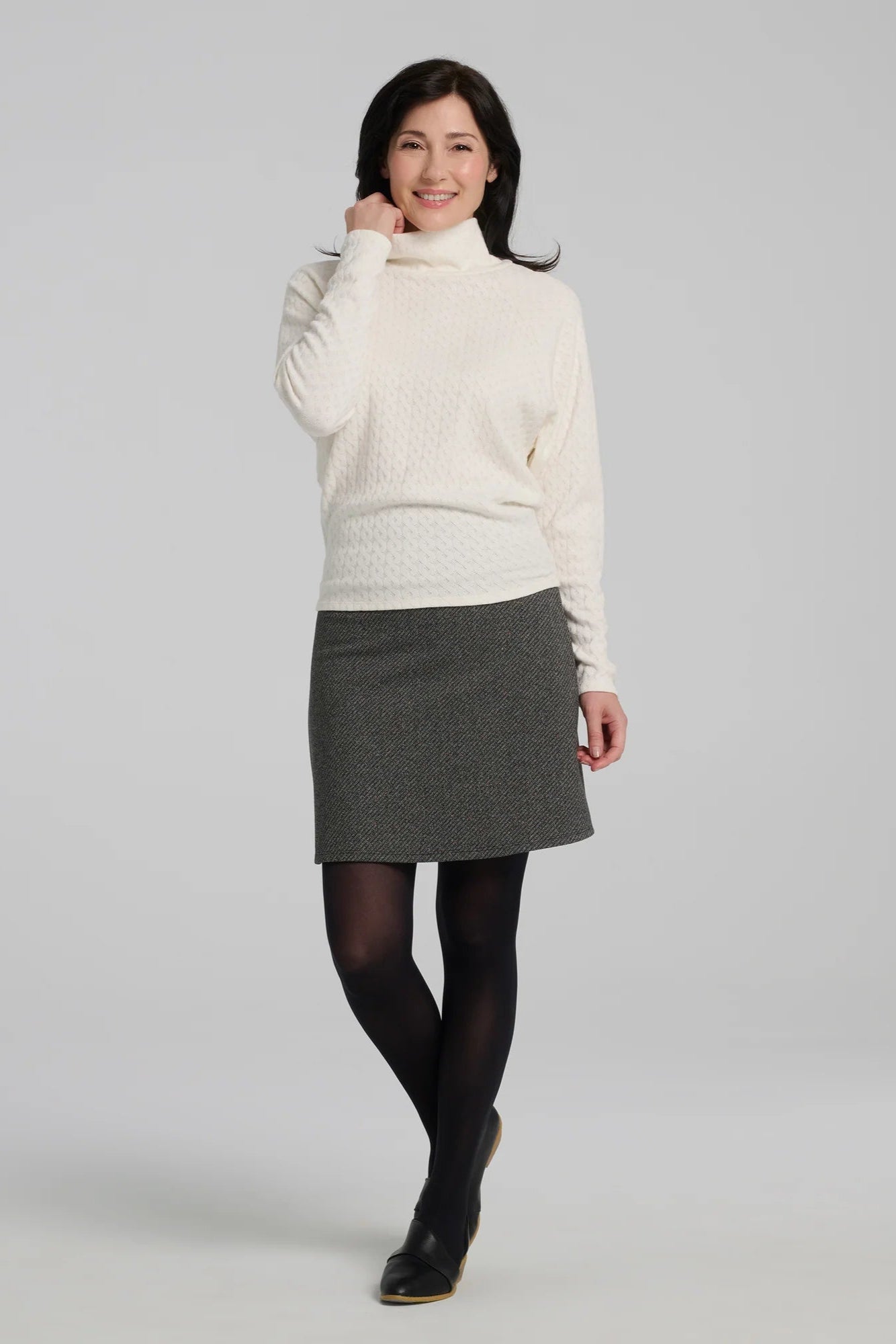 A woman wearing the Frost Skirt by Kollontai in Charcoal tweed, an above the knee length A-line skirt with an elastic waist.  She is wearing it with the Plume Top in and standing in front of a grey background. 