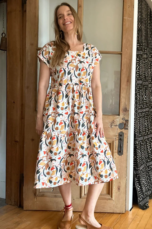 Woman wearing the Lavendula Dress by Kazak in Floral with cream, a short sleeved square neck midi dress with three ruffles. She is standing in a room with wooden floors and a door.