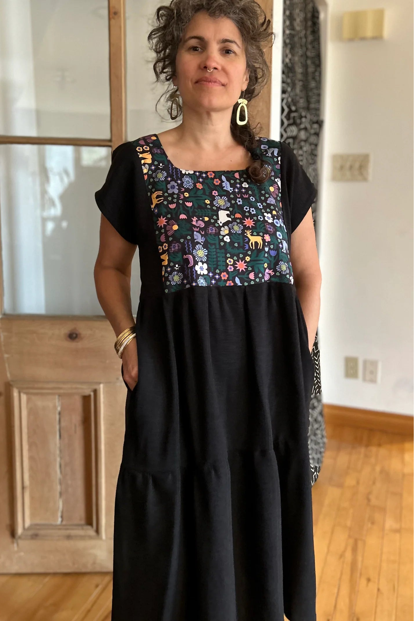 Woman wearing the Lavendula Dress by Kazak in Farmland, a short sleeved square neck midi dress with three ruffles. She is standing in a room with wooden floors and a door