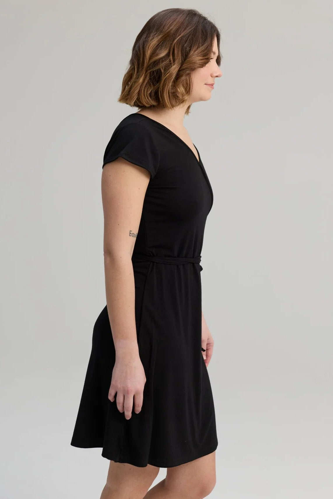 Side view of a woman wearing the Libellule Dress by Message Factory in Black, a short sleeved wrap dress that falls to the knee. She is standing against a plain background. 