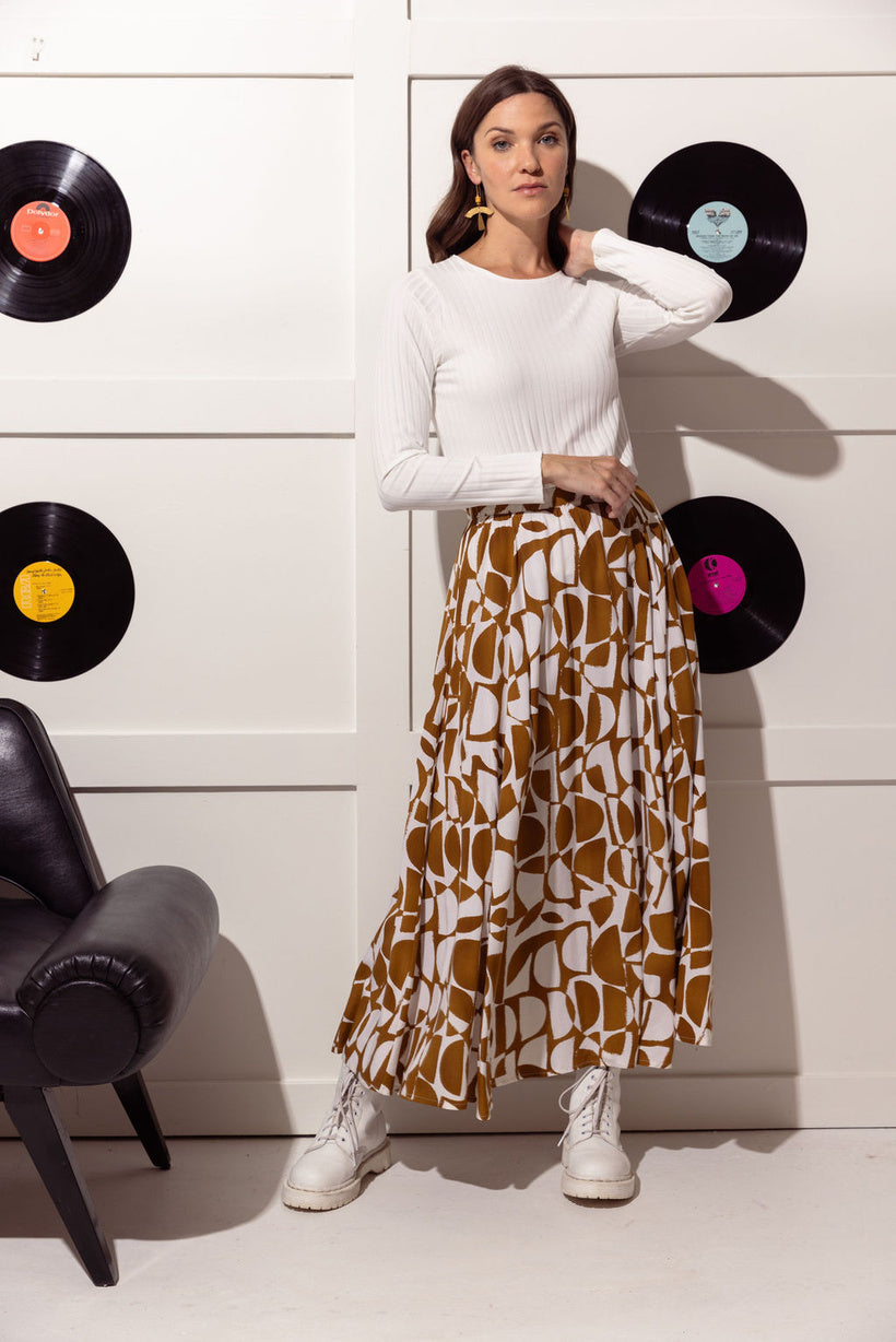 A woman wearing the long, pleated Hendrix Skirt by Cherry Bobin in Caramel and Cream. The skirt has an elastic waist and a semi-circle pattern.  The woman is wearing it with a white shirt, and is standing in front of a white wall with vinyl records on it  