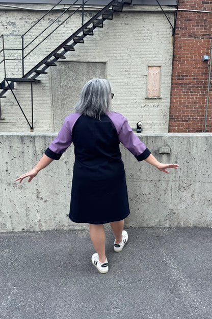 Back view of a woman wearing the Lorraine Dress by Solomia in Avocado/Purple Navy, an A-line colour-blocked dress in stretch denim with 3/4 sleeves and a knee-length hem. She is standing outside in front of a cement wall. 