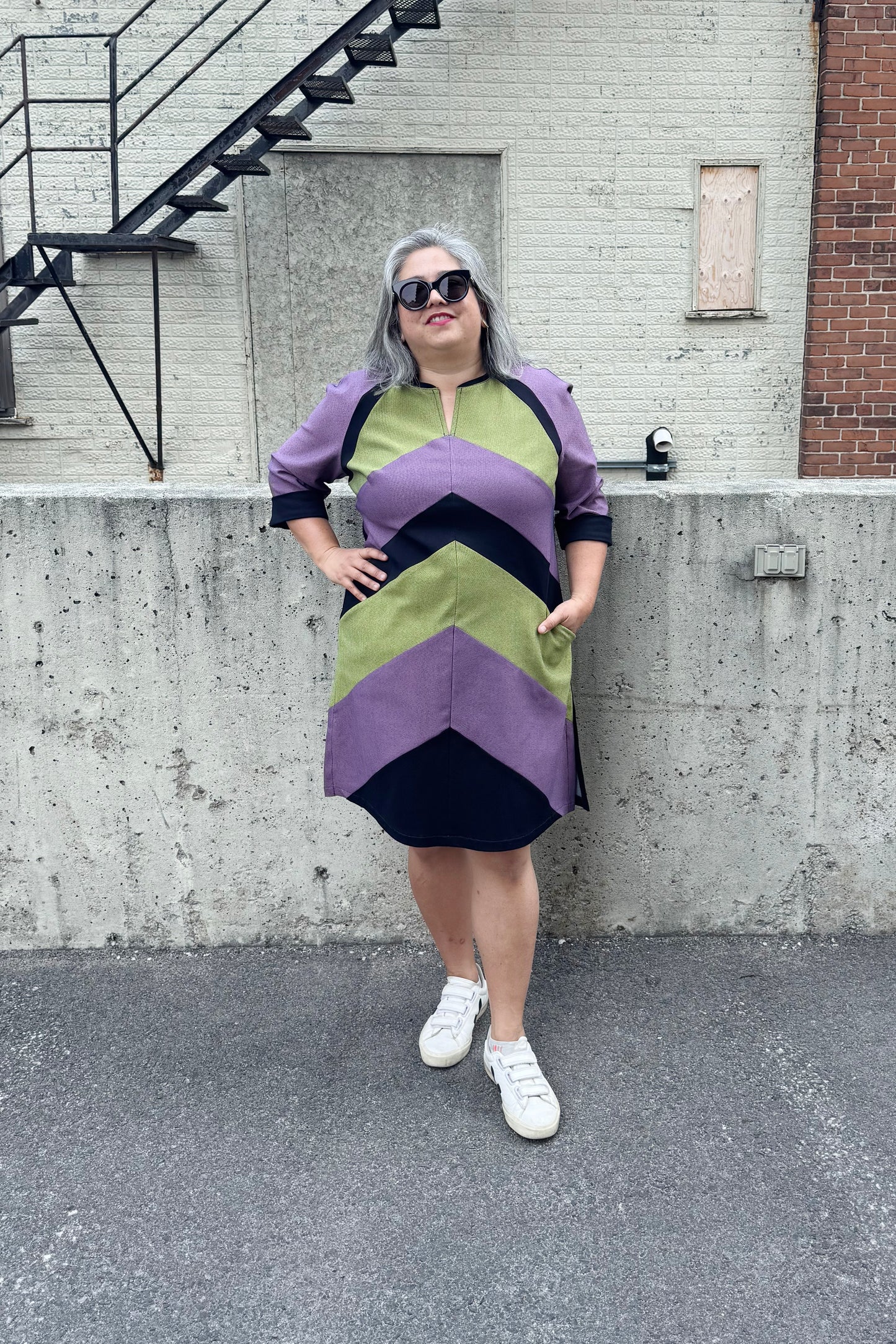 A woman wearing the Lorraine Dress by Solomia in Avocado/Purple Navy, an A-line colour-blocked dress in stretch denim with 3/4 sleeves and a knee-length hem. She is standing outside in front of a cement wall. 