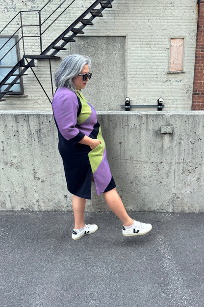 Side view of a woman wearing the Lorraine Dress by Solomia in Avocado/Purple Navy, an A-line colour-blocked dress in stretch denim with 3/4 sleeves and a knee-length hem. She is standing outside in front of a cement wall. 