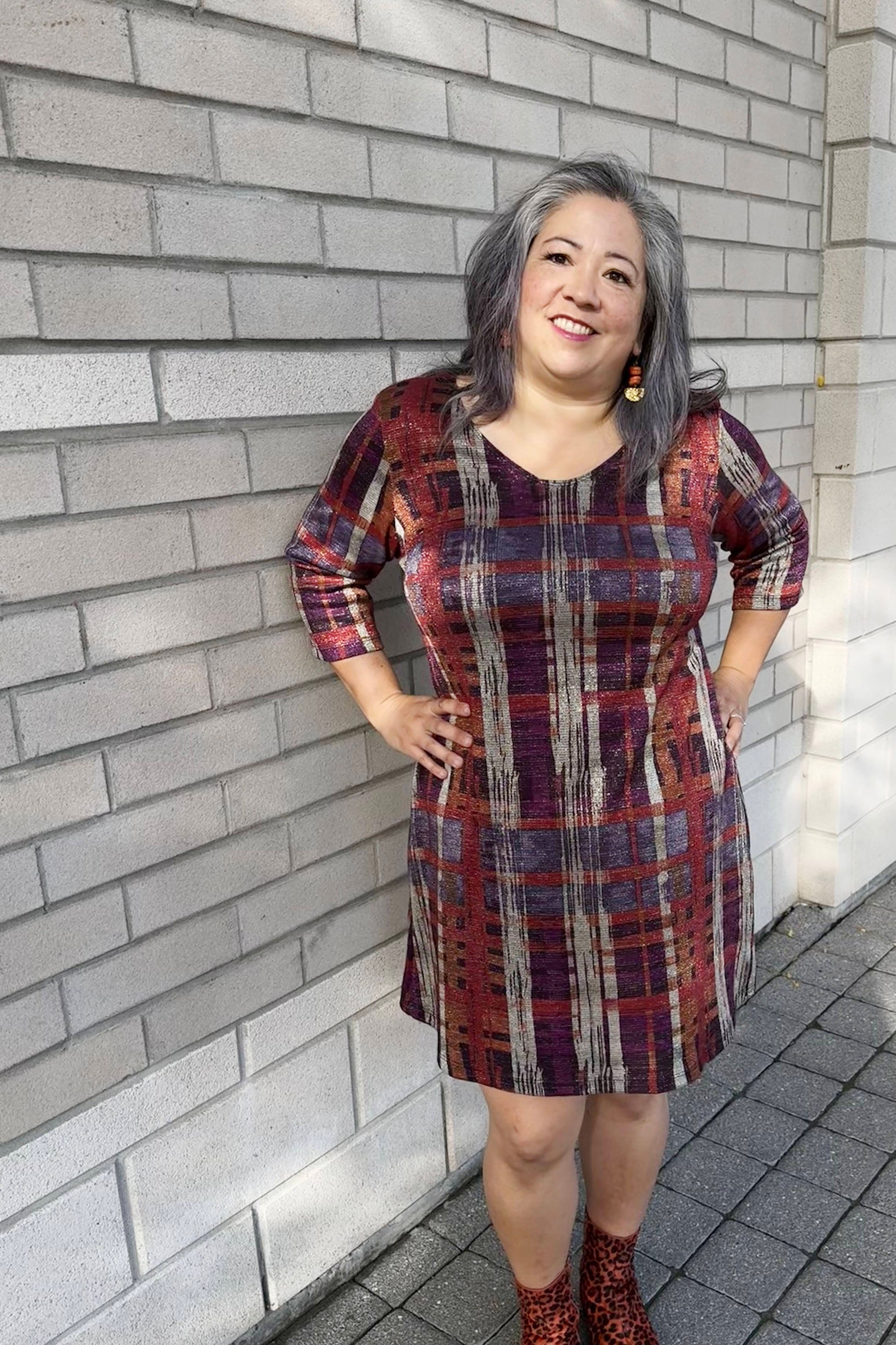 A woman wearing the Charlize Dress by Luc Fontaine in Multicolour Print in with a plaid effect in jewel tones with a touch of sparkle, featuring  a V-neck, 3/4 sleeves, and A-line shape that hits above the knee. She is standing outside in front of a brick wall. 