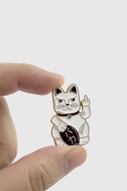 A hand holding the Lucky Cat Pin from Badaboöm Studio, a small enamel pin shaped like a fortune cat whose giving the finger, shown against a white background. 
