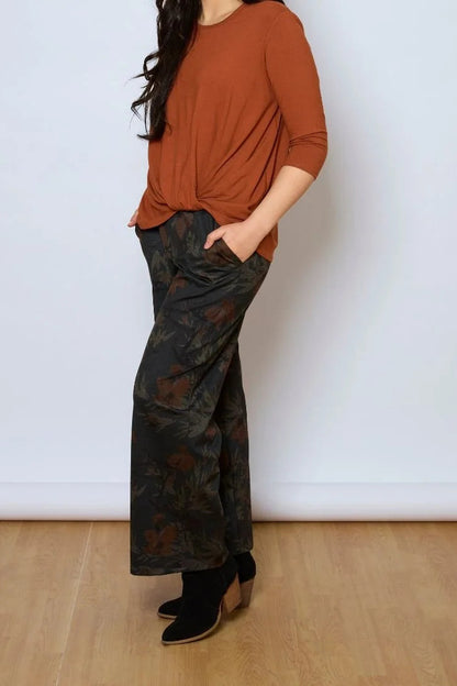 A woman wearing the Luisa Pants by Tangente in Floral, featuring an elastic waist with pleats, straight legs, and side pockets. She is wearing them with a rust coloured top and standing in front of a white background. 