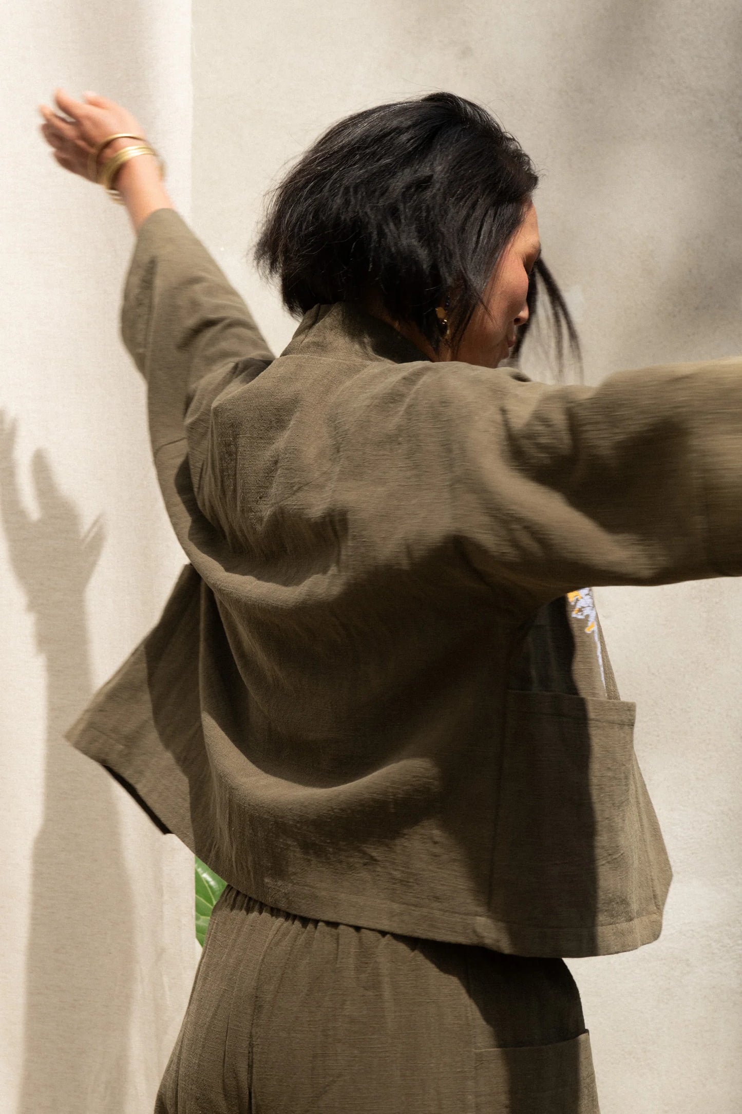 Back view of a woman wearing the Magnolia Screen-printed Jacket from Kazak in Olive with Dandelions, a short kimono style jacket with hand-screen printed dandelions, open front, 3/4-length sleeves, and big patch pockets. She is wearing it with matching pants and standing outside by a grey wall.