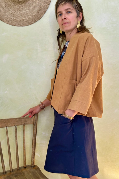 Side view of a woman wearing the Magnolia Jacket from Kazak in Turmeric, a kimono style jacket with 3/4 sleeves and large patch pockets. She is wearing it with a floral top navy and standing in a room with a wooden chair. 