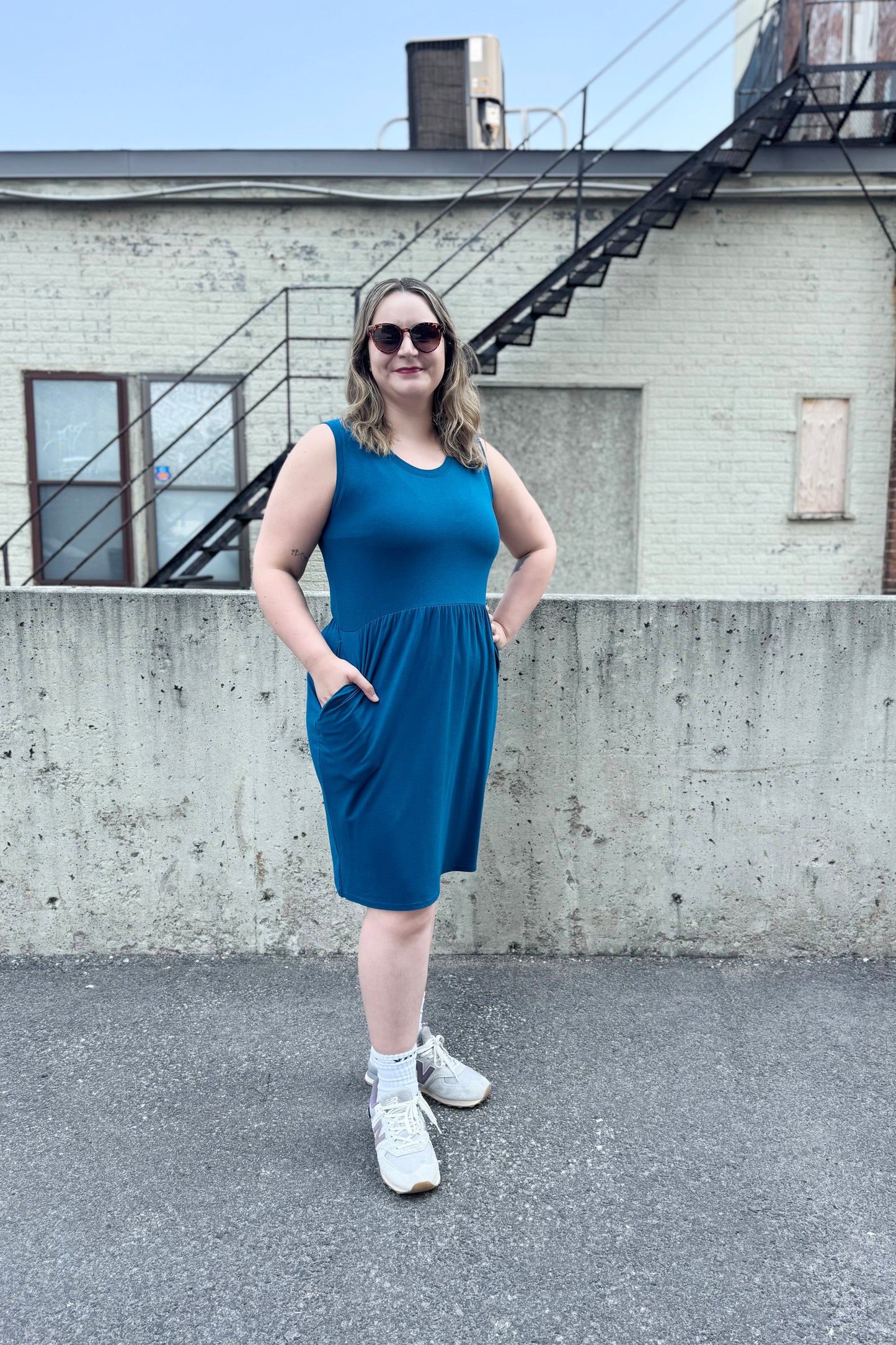 A woman wearing the Mai Dress by Tangente in Teal, a sleeveless bamboo jersey dress with a round neck and  gathered knee-length skirt. She is standing outside in front of a cement wall. 