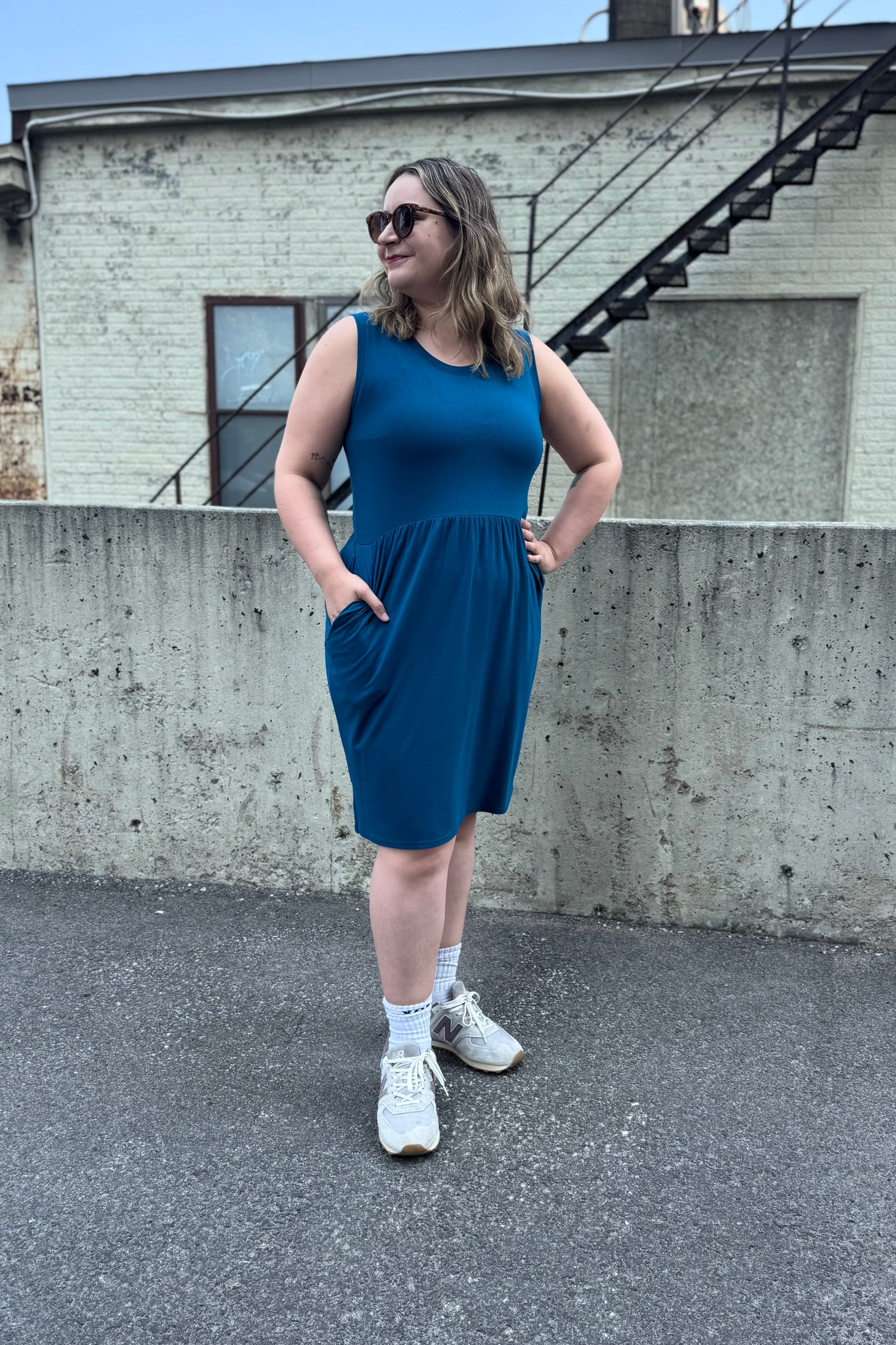 A woman wearing the Mai Dress by Tangente in Teal, a sleeveless bamboo jersey dress with a round neck and  gathered knee-length skirt. She is standing outside in front of a cement wall. 