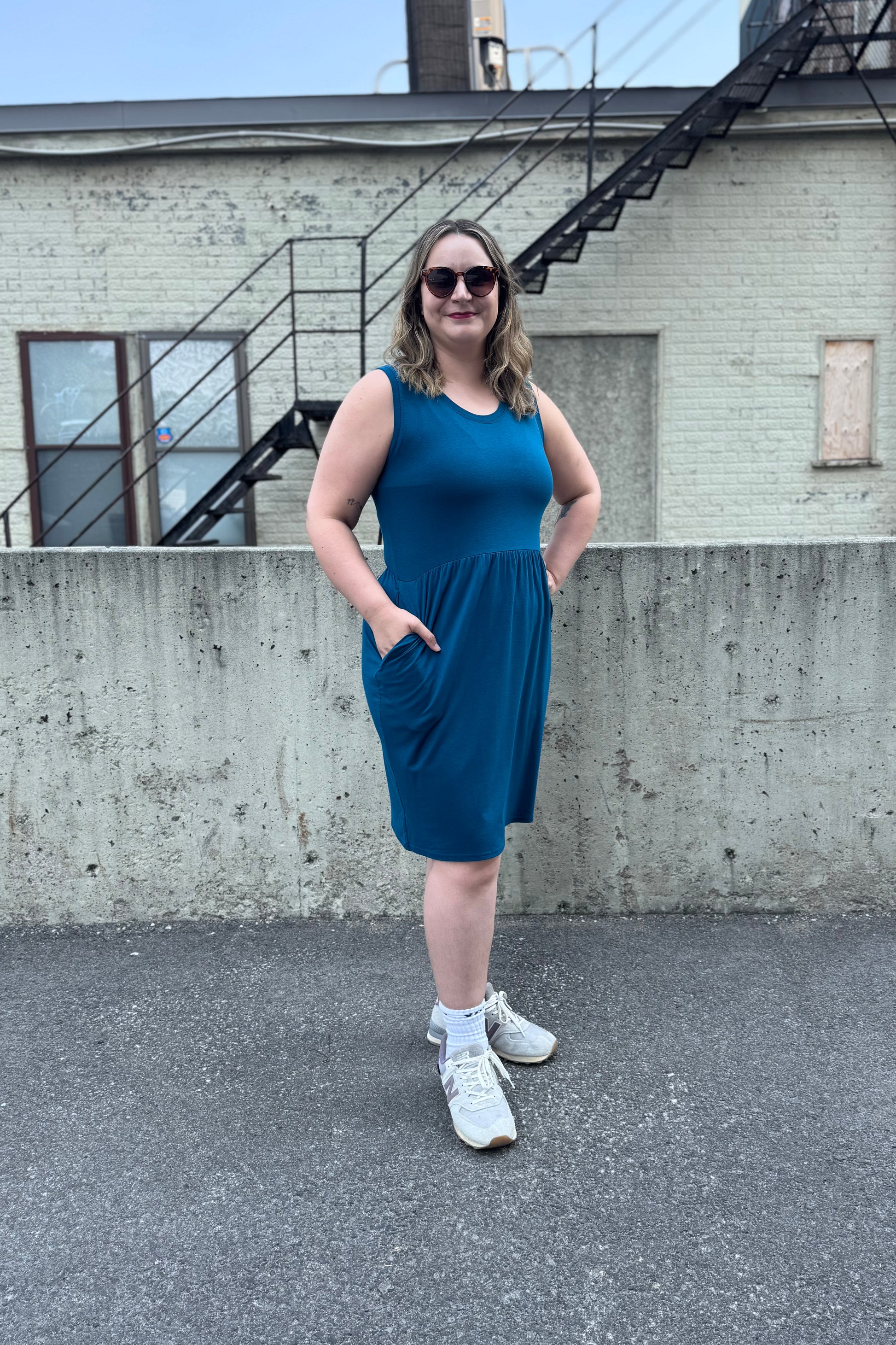 A woman wearing the Mai Dress by Tangente in Teal, a sleeveless bamboo jersey dress with a round neck and  gathered knee-length skirt. She is standing outside in front of a cement wall. 