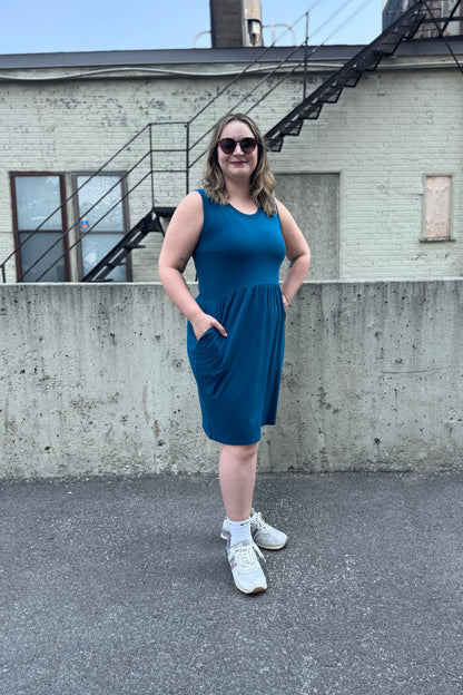 A woman wearing the Mai Dress by Tangente in Teal, a sleeveless bamboo jersey dress with a round neck and  gathered knee-length skirt. She is standing outside in front of a cement wall. 