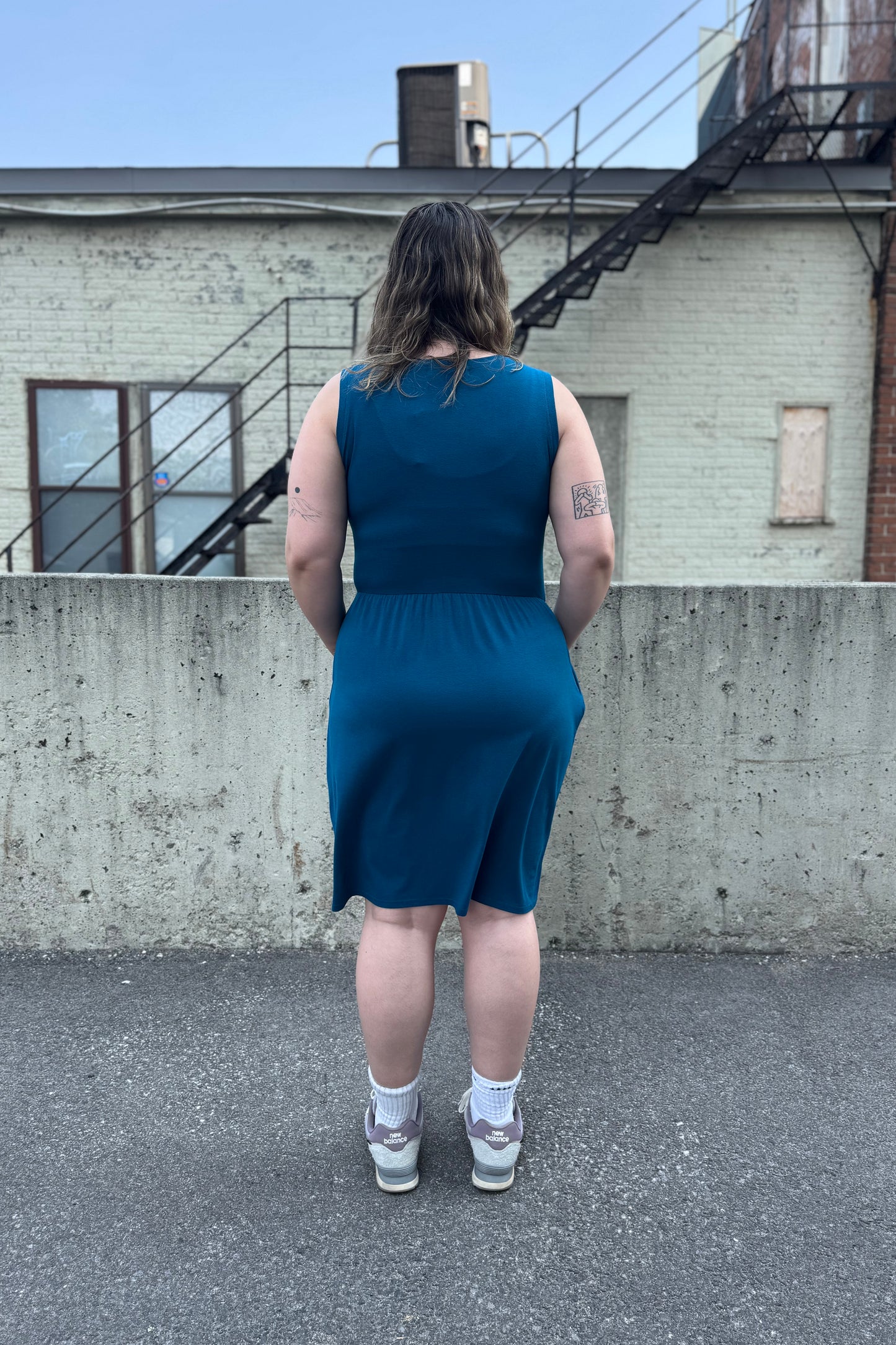 Back view of a woman wearing the Mai Dress by Tangente in Teal, a sleeveless bamboo jersey dress with a round neck and  gathered knee-length skirt. She is standing outside in front of a cement wall. 