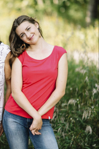 A woman wearing the Sol Top by Marie C in Coral, with a scoop neck and cap sleeves, with jeans, standing in a field. 